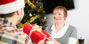 Image of older woman giving a red gift with a cream colored bow to a man in a santa hat in front of a Christmas tree