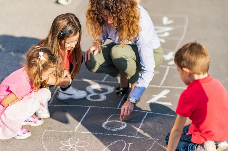 Daycare staff supervising young children playing outdoors during hot summer weather.