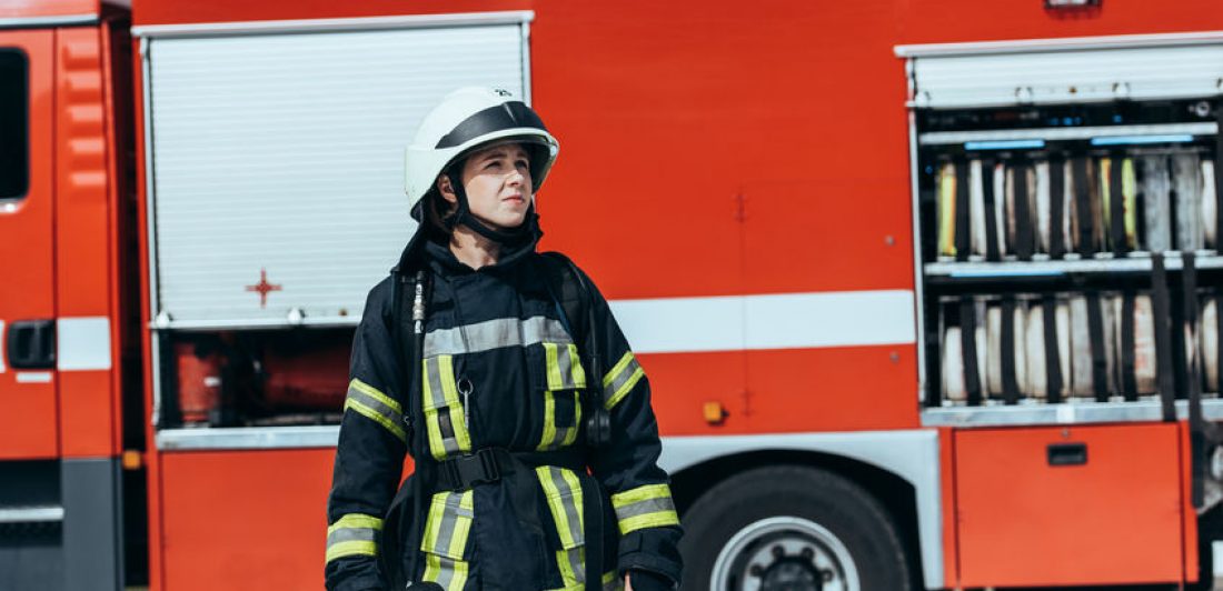 female firefighter in protective uniform standing on street with red fire truck behind Image of a female firefighter standing in front of a fire truck