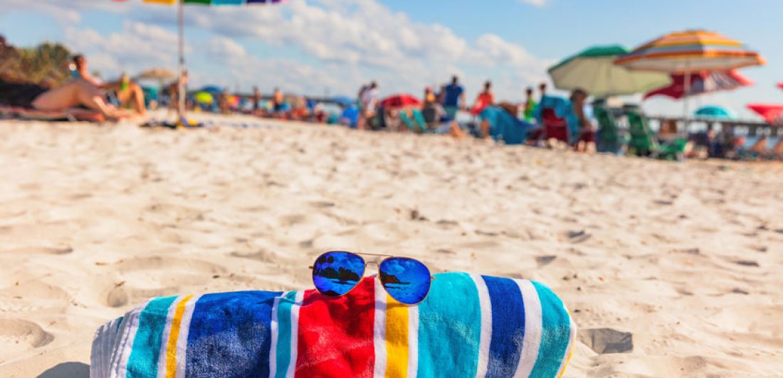 Beach towel and fashion sunglasses on Florida beach background -summer vacation travel concept copy space. Crowded popular USA destination for american holidays. Image of beach towel and sunglasses laying on the sand at a busy beach