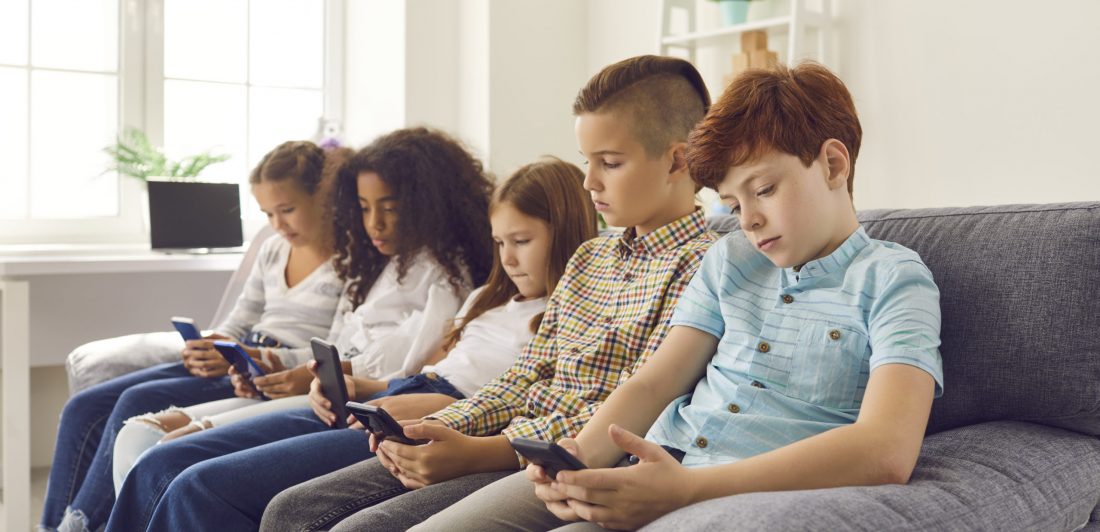 Group of multiethnic children sitting on sofa, using their smartphones and ignoring real life Image of children looking at their cell phones