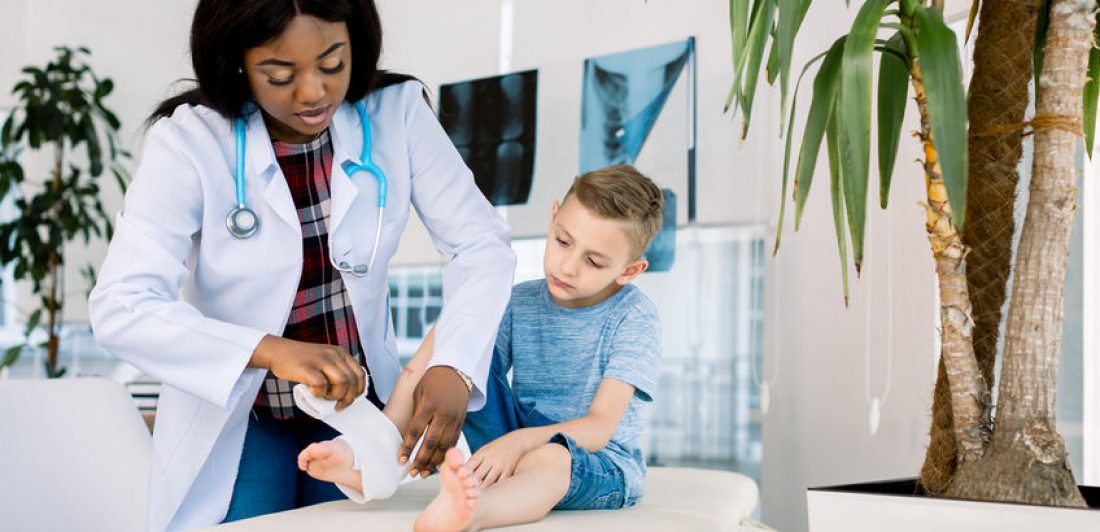 Putting bandage. Dark-skinned woman pediatrician wearing white coat putting bandage on leg of little boy Image of doctor wrapping a young boy's ankle