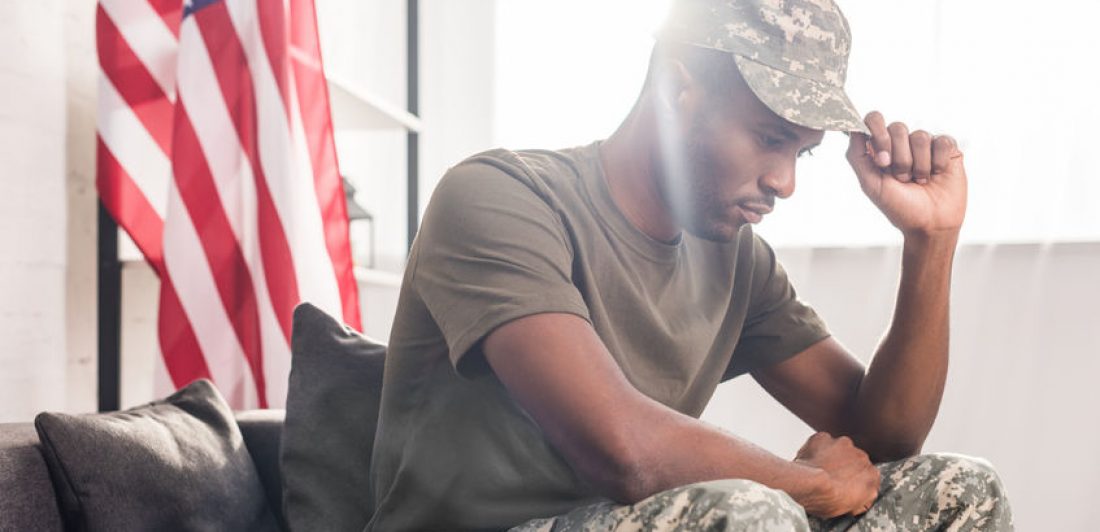 African american soldier in camouflage clothes sitting on sofa Image of sad man sitting in military uniform with flag in the background