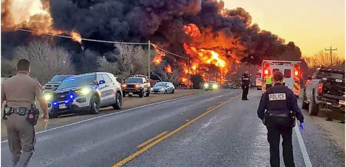 Image of a large fire started by an 18-wheeler and train collision with public safety personal parked and standing in the street