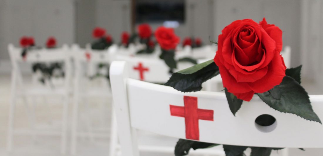 Image of white chairs with a white cross painted on the back and a red rose in each chair